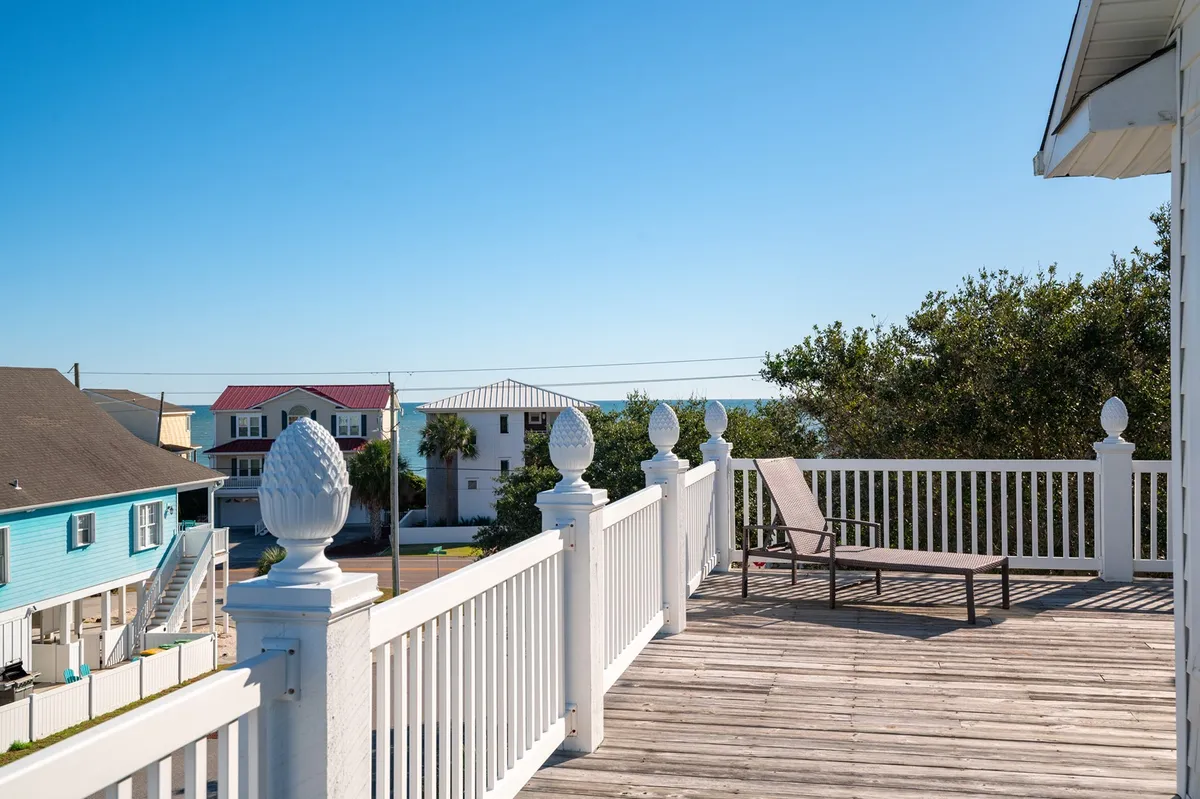 This exterior image captures a sun-drenched deck with white railings and a comfortable lounge chair, set against the backdrop of a blue house. The inviting outdoor space clearly hints at ocean views and the laid-back atmosphere of a beach community, echoing the promise of a relaxing getaway.