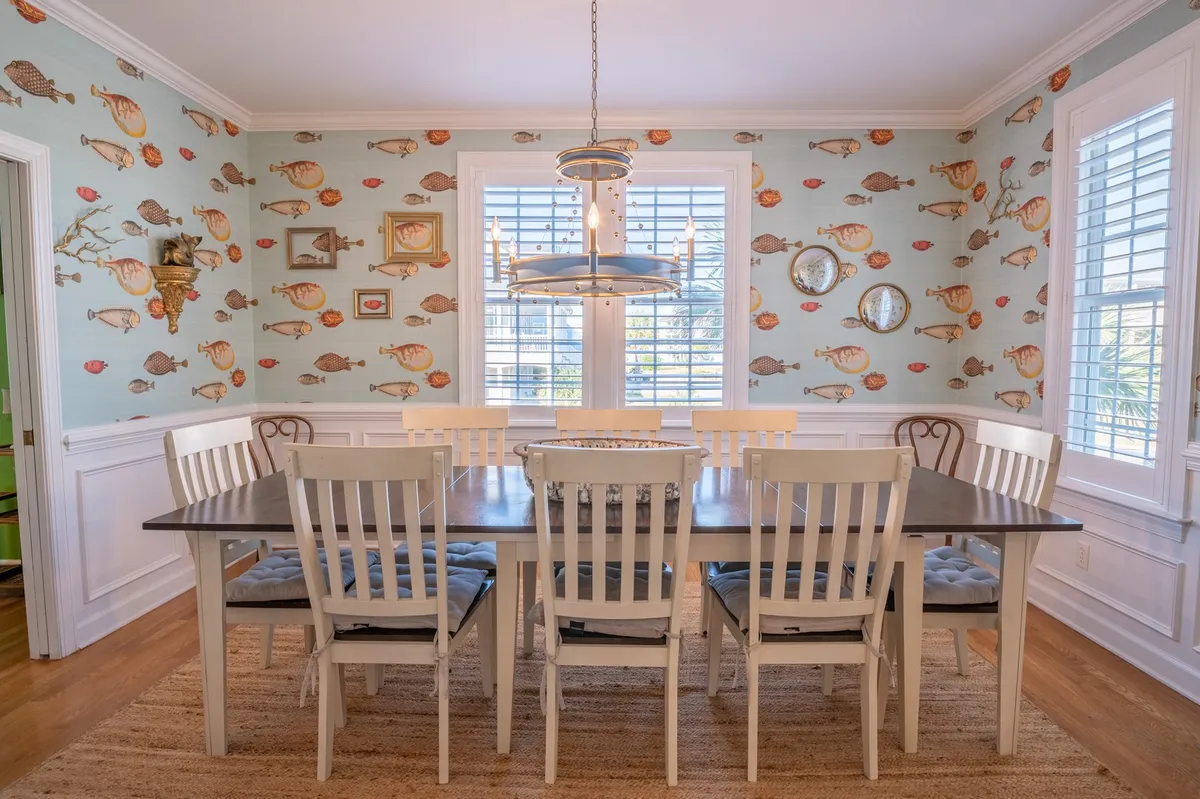 A large wooden dining table sits at the heart of the room, surrounded by windows with shutters that let in natural light. The walls are adorned with decorative fish wallpaper, and a modern light fixture adds a cheerful vibe, inviting groups to gather for meals and celebrations.