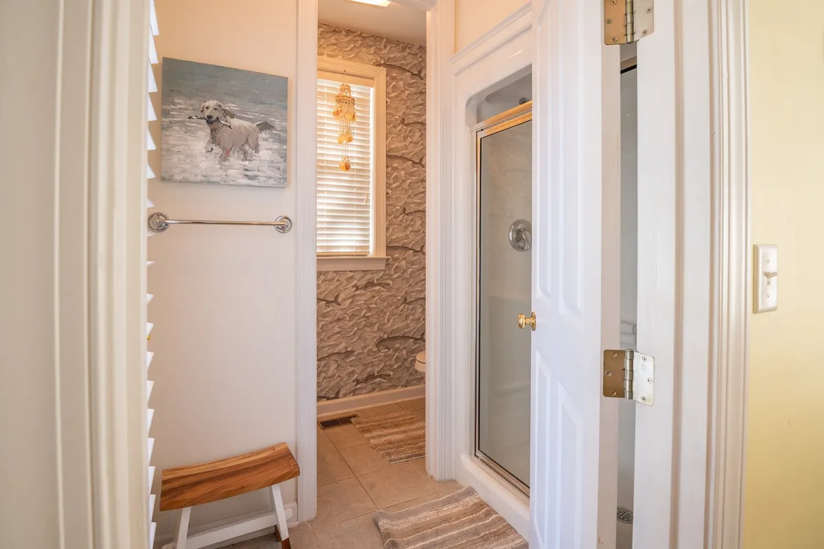 A serene, well-lit bathroom interior featuring a painting of a dog on the wall, a shower enclosure, a wooden bench, and a window letting in natural light.