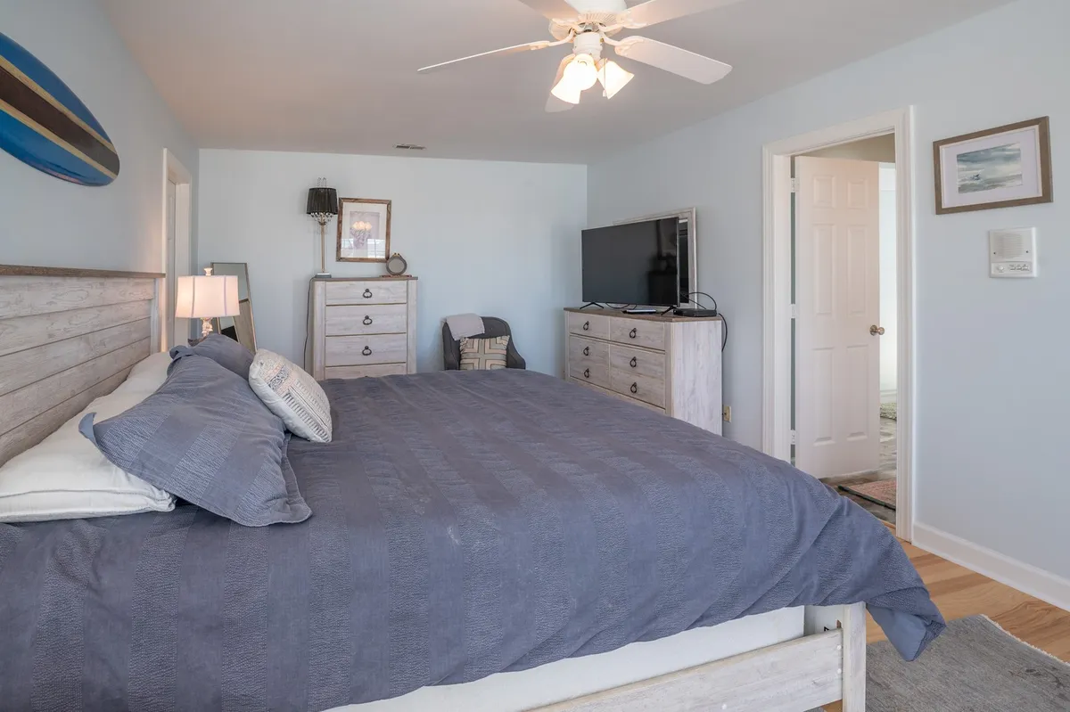 A calm and modern bedroom featuring a comfortable bed, a TV on the dresser, a lamp, and a surfboard accent. The room is styled for both relaxation and entertainment, offering a peaceful setting for restful nights or cozying up to watch TV.