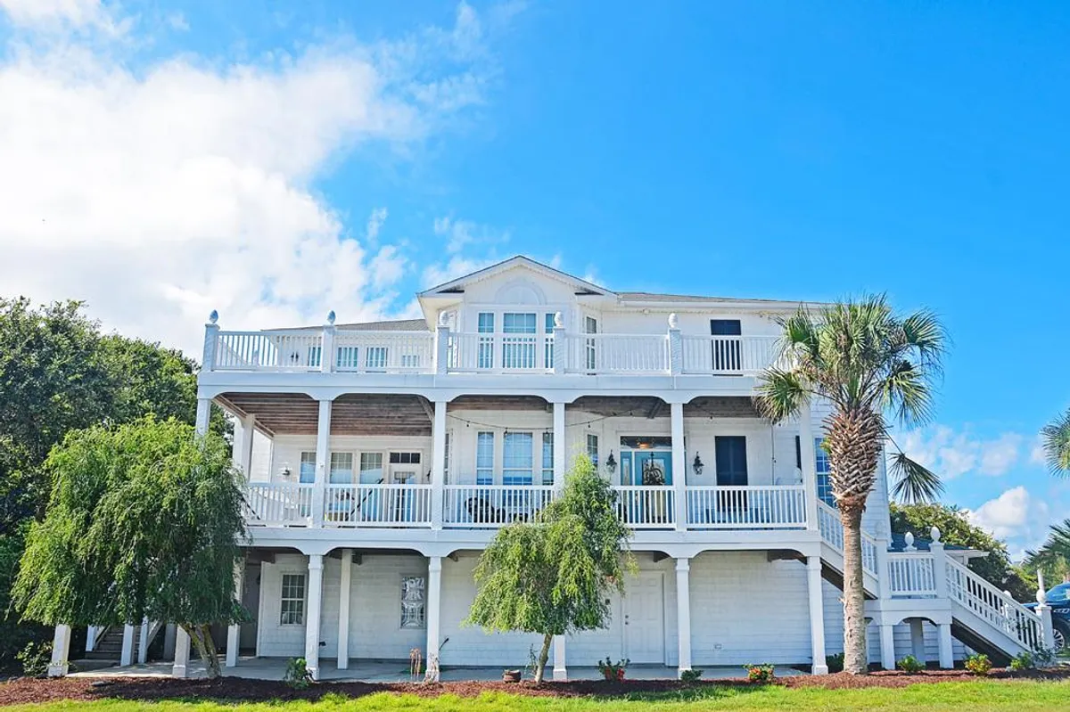 The image showcases the exterior of a grand single-family beach house at Kure Beach, featuring a spacious balcony with decorative railings framed by lush palm trees. The relaxed setting hints at opportunities for outdoor gatherings, enjoying morning coffee with ocean views, and taking in sunsets with family and friends.