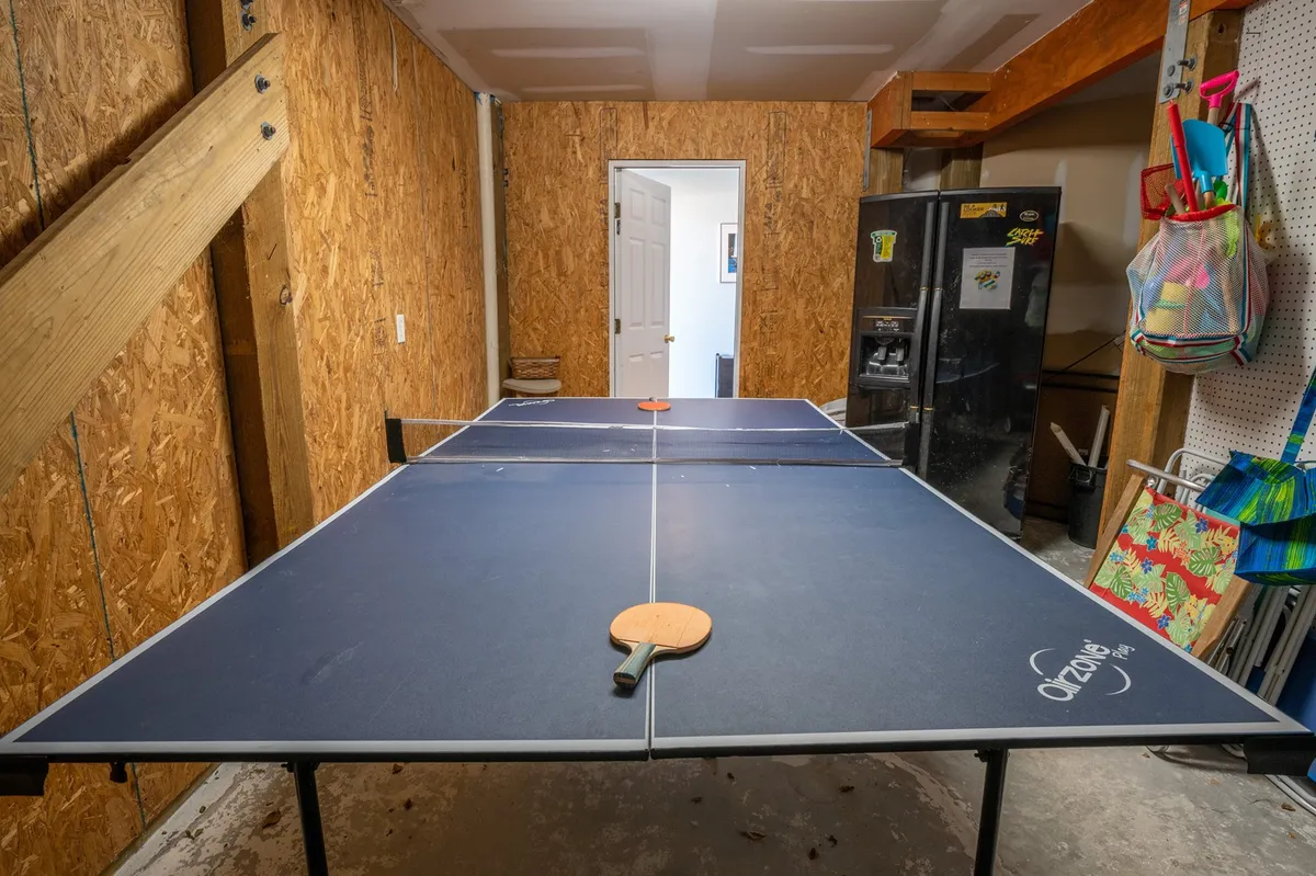 A lively playroom scene centered on a ping pong table, with a paddle resting on the surface, a refrigerator in the background for easy access to snacks and drinks, and colorful bags hanging on the wall. A doorway hints at the room’s connection to other spaces, creating a sense of openness ideal for active family gatherings and friendly tournaments.