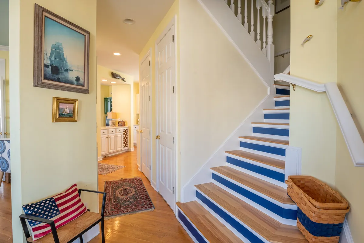 A sunlit staircase accented with blue, sporting inviting artwork and a striped chair with an American flag motif, creates a mood of hospitality and movement. The rug and open atmosphere reinforce a sense of welcome and anticipation.