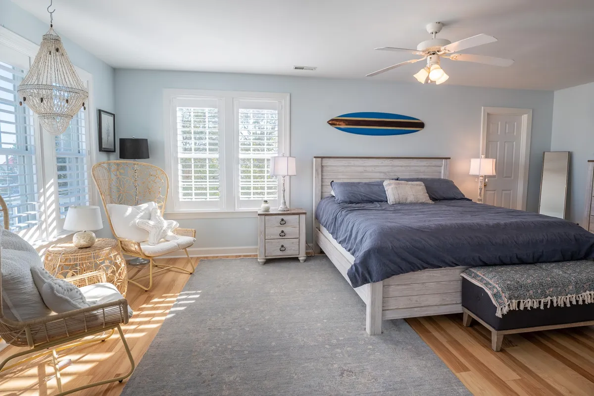A serene bedroom with a bed featuring crisp linens, a decorative surfboard on the wall, a cozy rattan chair by the window, and nightstands with lamps. Natural light streams into the room, creating a tranquil atmosphere ideal for unwinding after a day at the beach.