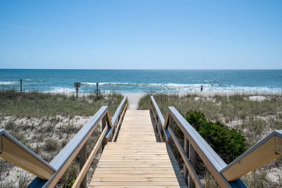 This image showcases a wooden walkway lined with dune grass leading directly to a wide, sandy beach, with a view of the ocean and the horizon beyond. The sunset sky casts a golden glow, inviting families to head out for a stroll, swim, or beachside play. The scene captures the private and easy beach access highlighted in the section.