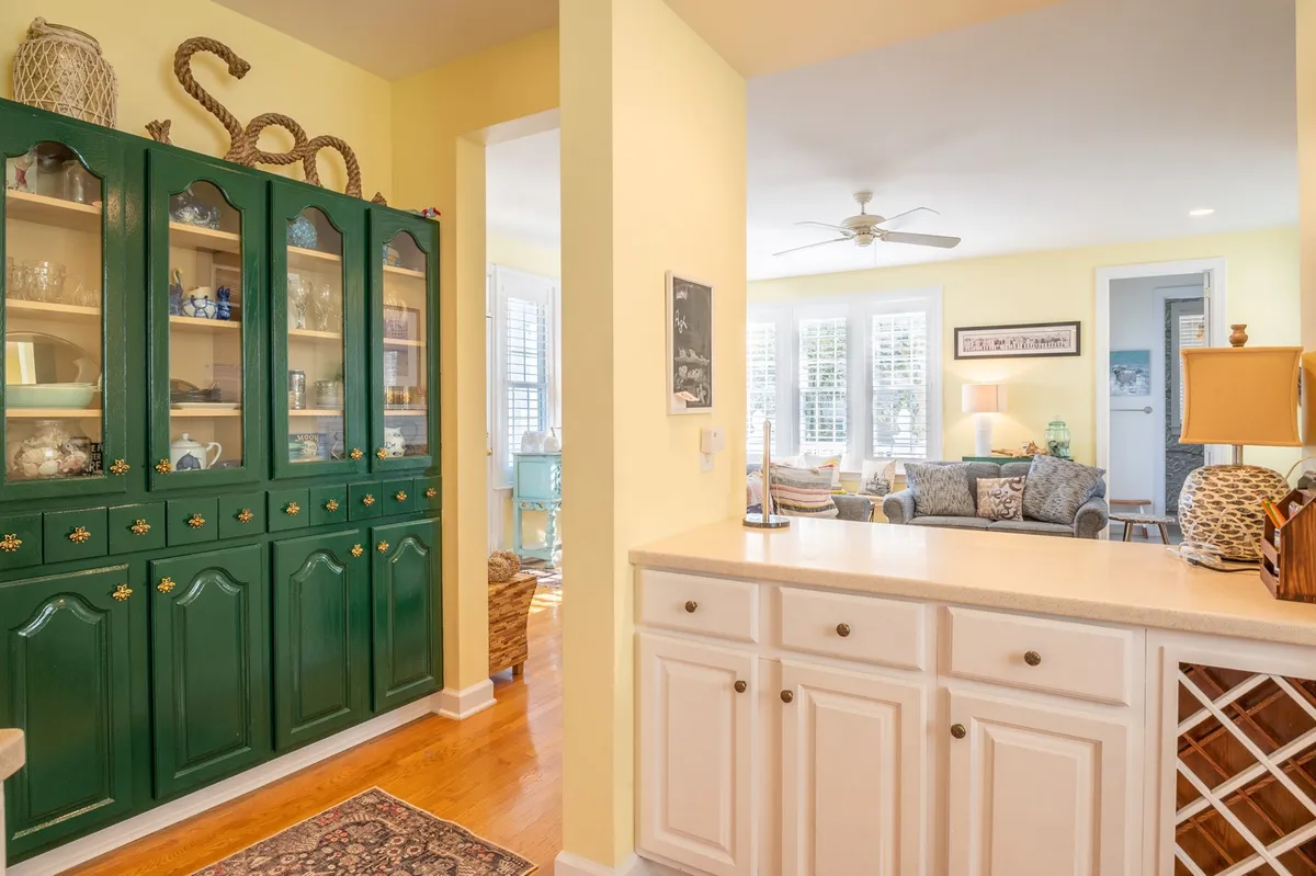 A sunlit open-plan space featuring a farmhouse kitchen with a green display cabinet and a spacious kitchen island. In the background, the inviting living area is visible, seamlessly connecting kitchen, dining, and lounging zones. The space feels vibrant and perfect for entertaining or family gatherings.