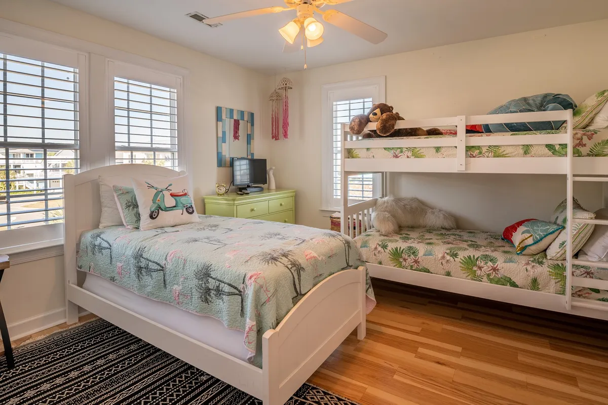 A playful, cozy bedroom designed for children, featuring a colorful bunk bed, a single bed with a vibrant quilt, a green dresser, and large windows that let in ample daylight. The space feels inviting and filled with opportunities for both rest and play.