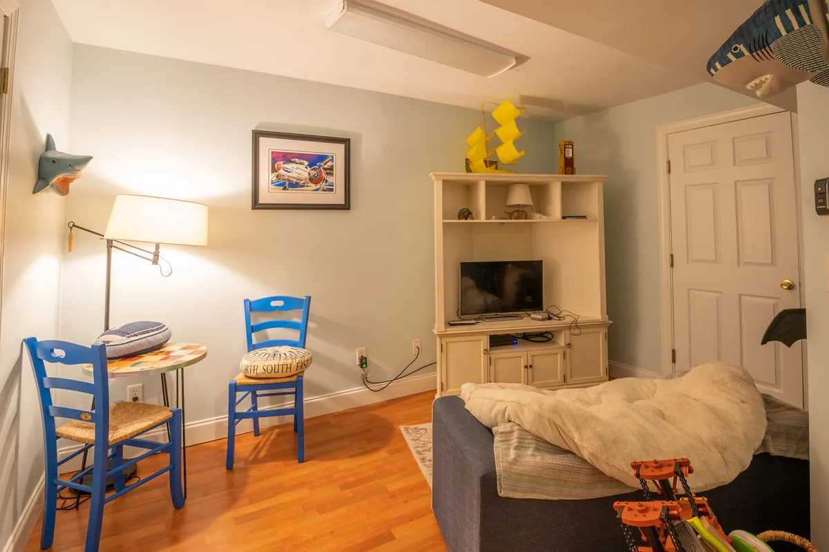 A cozy, well-lit interior with blue chairs, framed artwork, a TV unit, and a playful shark decoration. The space is inviting for both socializing and creative play, hinting at activities like movie nights, reading, and kid-friendly games.