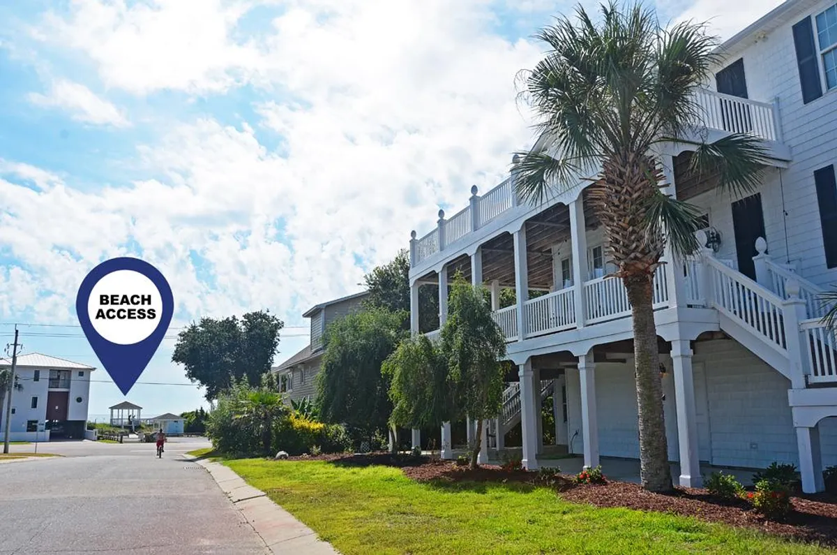 This exterior image features a 'beach access' sign, palm trees, and the exterior of the building, inviting viewers to envision themselves heading out for a seaside stroll. The lawn area and visible porch evoke a sense of easy outdoor living and provide a perfect setting for guests with pets.