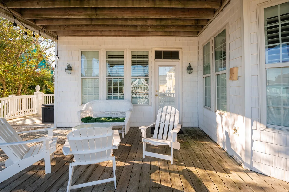 A tranquil deck setup with a white wicker couch, adirondack chairs, and a wooden deck, offering a serene spot for group relaxation. The doors leading inside suggest fluid movement between communal indoor and outdoor spaces.